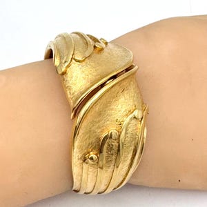 May include: A gold-toned cuff bracelet with a textured surface and intricate design. The bracelet features a curved shape with detailed embellishments, creating a bold and elegant statement piece. The bracelet is worn on a wrist.