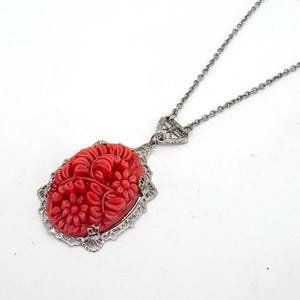 May include: A silver-toned necklace featuring a red oval pendant with a carved floral design. The pendant is framed by a decorative silver border and hangs from a delicate chain. The necklace is set against a white background.