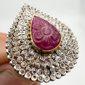 May include: A pear-shaped ruby ring with a carved floral design, surrounded by a halo of small diamonds.