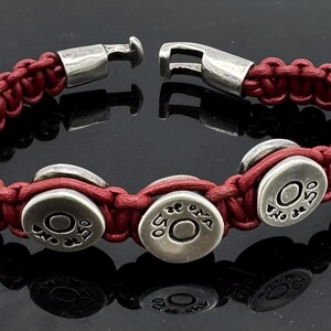 May include: A red leather bracelet with silver metal accents. The bracelet features four round silver charms with the text "No De 50" engraved on each.