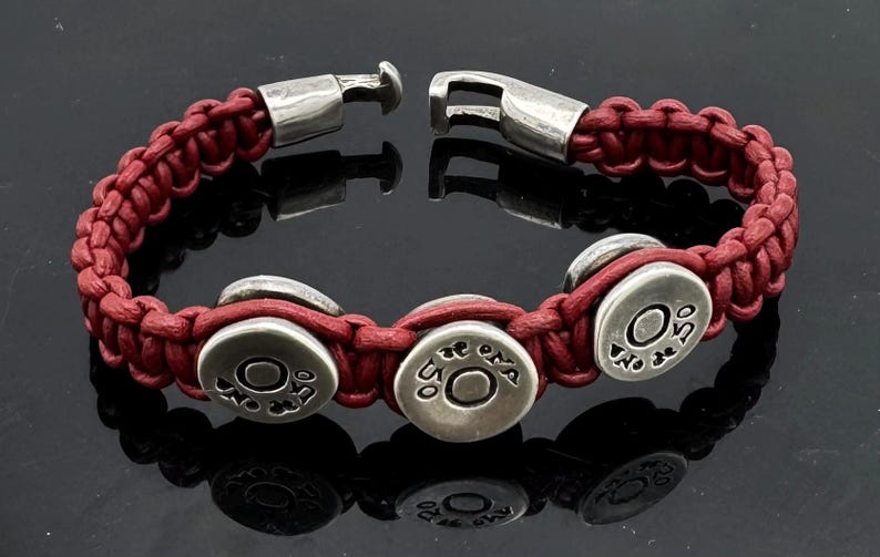 May include: A red leather bracelet with silver metal accents. The bracelet features four round silver charms with the text "No De 50" engraved on each.