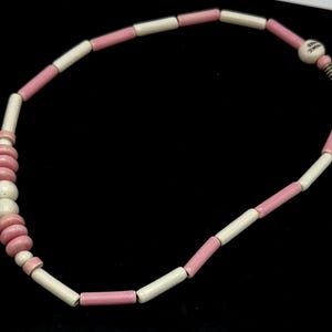 May include: A necklace featuring alternating pink and off-white cylindrical beads. The necklace has a cluster of pink and off-white round beads on one side. The clasp has the text "MADE IN JAPAN".