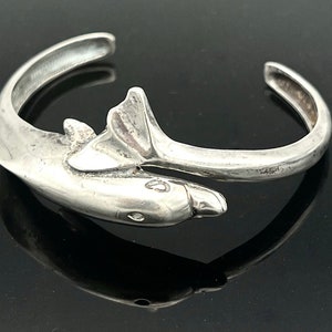 May include: A silver-toned cuff bracelet featuring a dolphin design. The bracelet wraps around the wrist, with the dolphin's body forming the main part of the cuff. The dolphin has a detailed head and tail, and the bracelet is approximately 2.5 inches in diameter.