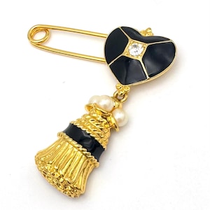 May include: Gold-tone safety pin brooch with a black heart-shaped top, a central clear rhinestone, and a tassel pendant. The tassel is gold with black accents and faux pearls. A unique and elegant accessory.