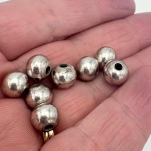 May include: A selection of silver beads with a shiny finish. The beads are spherical with a hole through the middle, and are likely used for jewellery making or crafts. The beads are shown in a hand.