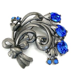 May include: A silver tone brooch with a floral design and blue rhinestones. The brooch features a bow at the bottom and a cluster of flowers with blue rhinestones at the top.