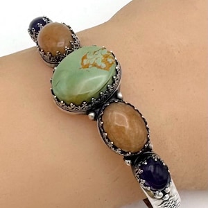 May include: Silver bracelet with five oval gemstones in green, brown, and purple. The stones are set in ornate silver settings. The bracelet is a hinged cuff style.
