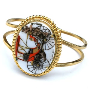 May include: Gold-coloured cuff bracelet with an oval centrepiece depicting a vintage bicycle in red, black, and white. The bracelet has a textured band and a beaded border. The text "Durgea 1893" is visible on the centrepiece.
