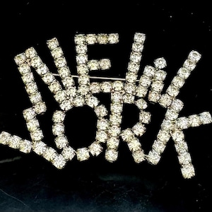 以下が含まれることがあります： 「NEW YORK」の文字をスタイリッシュなフォントで綴った、キラキラ光るラインストーンのブローチ。文字は透明でファセットカットされた石で密に詰め込まれており、黒い背景にきらめく効果を生み出しています。ブローチはシルバーカラーのピンバックが付いています。