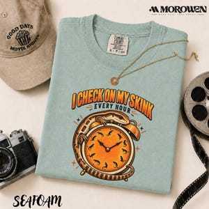 May include: A light green t-shirt featuring a graphic of a skink lizard wrapped around an orange alarm clock. The text reads I CHECK ON MY SKINK EVERY HOUR. The clock face has small lizard silhouettes instead of numbers. A gold chain necklace rests on the shirt.