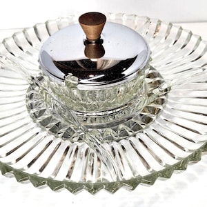 May include: A clear glass serving dish with a circular, ribbed design and a scalloped edge. Centered on the dish is a small glass bowl with a polished chrome lid, featuring a dark brown wooden knob handle and a matching clear glass serving spoon.