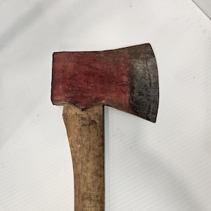 May include: A single-bit axe featuring a weathered wooden handle and a metal head with a dark, oxidized cutting edge and a faded red painted finish on the poll and cheeks, showing signs of wear and texture across the entire tool surface.