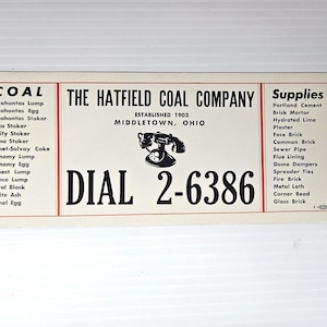 May include: A rectangular white sign with black text featuring The Hatfield Coal Company, established 1903 in Middletown, Ohio. It lists coal types and supplies, displays a black rotary telephone graphic, and includes the phone number DIAL 2-6386.