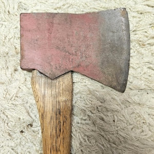 May include: A hand axe featuring a weathered, dull red metal head with visible signs of wear and oxidation, securely attached to a light brown wooden handle with a prominent vertical grain pattern, showing a sturdy construction and functional design.