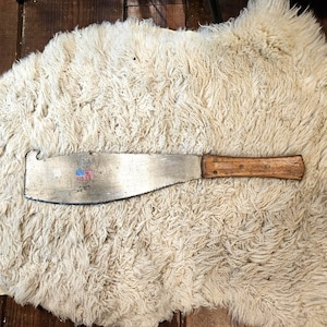 May include: A metal machete with a wooden handle secured by two rivets. The blade features a small hook at the top edge and a printed graphic of the United States flag with the text USA. The blade surface shows signs of wear and metallic texture.