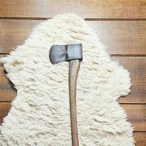 May include: A hand axe featuring a dark, weathered metal head with a slightly curved cutting edge and a long, straight wooden handle. The tool displays a worn, textured appearance on the metal surface, contrasting with the smooth, grain-patterned wood of the handle, which extends vertically through the center of the frame.