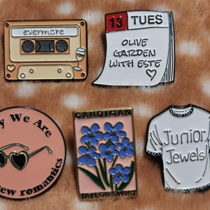 Op de afbeelding: Vijf emaille pins: een beige cassettebandje met evermore, een wit kalenderblad met 13 TUES OLIVE GARDEN WITH ESTE, een roze cirkel met hartvormige zonnebril, een rechthoekige kaart met blauwe bloemen en een wit t-shirt met de tekst Junior Jewels.