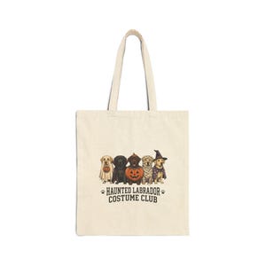 Peut inclure: Un sac fourre-tout en toile beige avec une illustration de cinq chiens Labrador en costumes d'Halloween, incluant un fantôme, un vampire, une citrouille, une momie et une sorcière. Sous les chiens, le texte indique HAUNTED LABRADOR COSTUME CLUB.