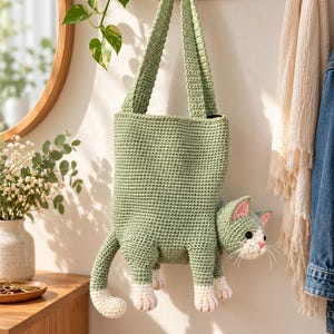 Puede incluir: Un bolso tote de ganchillo en color verde salvia con forma de gato. El bolso cuenta con una correa larga, un cuerpo rectangular y una cabeza, cola y cuatro patas de gato tridimensionales que sobresalen. Tiene patas blancas y orejas rosas.
