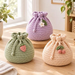 May include: Three round, crocheted drawstring pouches in light green, lavender, and pale peach. The green pouch features a small strawberry applique, the lavender one has a white flower with a green leaf, and the peach one displays two small cherries.