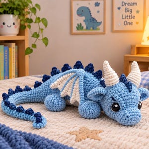 Puede incluir: Un dragón de peluche tejido a ganchillo en color azul claro con espinas azul oscuro a lo largo de la espalda y la cola, alas blancas y dos cuernos blancos. El dragón tiene grandes ojos de seguridad negros sobre una manta color crema con estrellas.