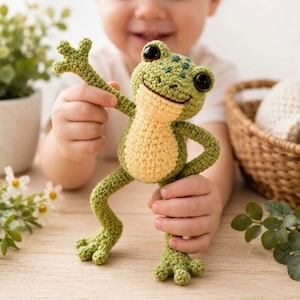 Könnte beinhalten: Ein gehäkeltes Froschspielzeug mit hellgrünem Körper, hellgelbem Bauch, dunkelgrünen Punkten auf dem Kopf und großen schwarzen Augen. Der Frosch hat vier Gliedmaßen, wobei ein Arm erhoben ist und der andere an der Seite ruht, gefertigt aus Garn.