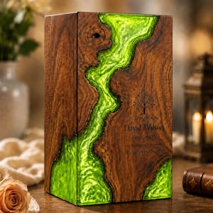 Könnte beinhalten: Eine rechteckige Urne aus Holz mit einer zentralen, flussartigen Einlage aus durchscheinendem, hellgrünem Harz. Das Holz hat eine dunkelbraune Maserung und die Vorderseite ist mit einem Baum-Symbol sowie dem Text In Loving Memory, David Wilson, 1938-2022 und Always in our hearts graviert.