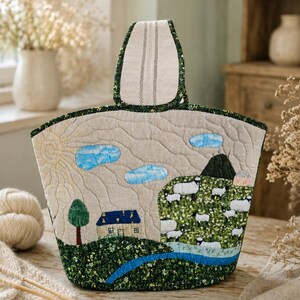 May include: A quilted fabric bag featuring a pastoral landscape scene with a house, a green hill, and a flock of sheep. The design includes a yellow sun, blue clouds, a green tree, and a blue stream. The bag has a single top handle and beige fabric.