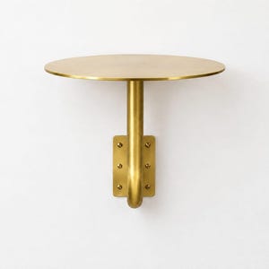 Puede incluir: Un estante de pared con una superficie circular de latón sostenida por un poste cilíndrico vertical. El poste se conecta a una placa de montaje rectangular de latón asegurada por seis tornillos visibles, todo con un acabado en tono dorado.