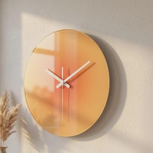 May include: A circular wall clock featuring a smooth, gradient face that transitions from soft yellow to warm orange tones. The clock has three thin, white hands positioned at the centre, with no visible numbers or markings on the glass surface.