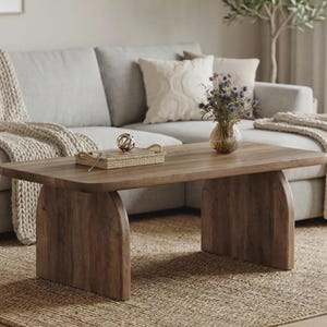 Puede incluir: Una mesa de centro rectangular de madera con una superficie superior lisa y plana y dos soportes laterales robustos en forma de arco. La mesa está hecha de madera de tono medio con un patrón de veta visible, proporcionando una base estable.