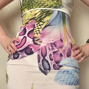 May include: A sleeveless dress featuring a white base with a vibrant, multi-coloured graphic print. The design includes large, detailed patterns of green pineapple textures, purple and black snake scales, and blue and pink floral elements.