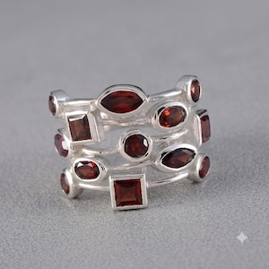 May include: A multi-band silver ring featuring a collection of deep red garnet gemstones in various shapes, including marquise, square, and round cuts, set within a polished silver frame that creates a layered and openwork appearance.