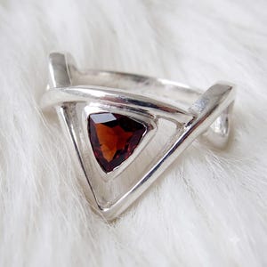 May include: A silver ring featuring a triangular-cut deep red gemstone set within a geometric frame. The band consists of two intersecting silver bars that form a triangular shape around the central stone, creating a layered and open design.