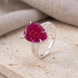 Op de afbeelding: Een zilveren ring met een grote, peervormige, diep magentakleurige edelsteen met een textuurrijk, kristallijn oppervlak. De ringband is gemaakt van gepolijst zilver en bevat gedetailleerde kraalversieringen langs de zijkanten.