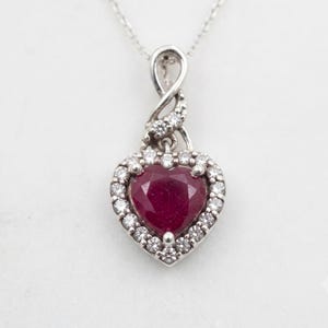 May include: A silver-toned necklace featuring a heart-shaped red gemstone pendant surrounded by a halo of small clear stones. The pendant is suspended from a twisted silver-toned bail that is also accented with small clear stones on a chain.