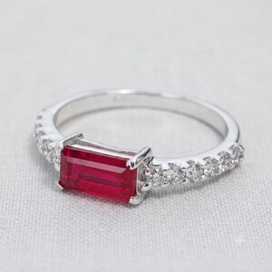 Può includere: Un anello color argento con una pietra preziosa rossa rettangolare a taglio baguette al centro, affiancata da una fila di piccole pietre chiare a taglio rotondo incastonate lungo la fascia su entrambi i lati, per un aspetto simmetrico.