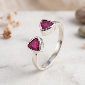 May include: A silver open-style ring featuring two triangular-shaped, deep pink gemstones set at each end of the band. The metal band has a smooth, polished finish, and the two stones are positioned to face each other with a gap between them.
