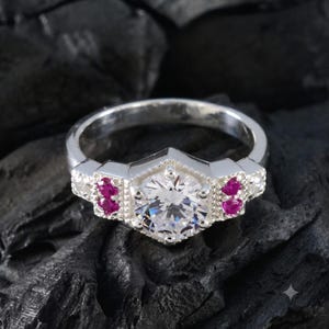 Peut inclure: Une bague argentée ornée d'une grande pierre centrale ronde et transparente à facettes, sertie dans une monture hexagonale. L'anneau est agrémenté de chaque côté de deux petites pierres rondes rose vif et de détails métalliques perlés.