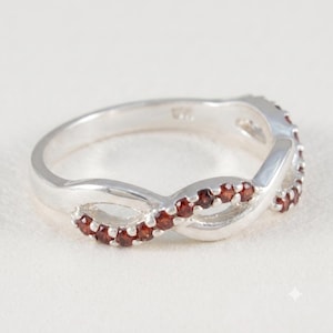 Può includere: Un anello in argento con un design a fascia intrecciata a forma di infinito. La superficie della fascia è incastonata con una fila di piccole pietre preziose granato rosso scuro, rotonde, che seguono la curva del metallo con brillantezza.