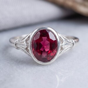 May include: A silver ring featuring a large, oval-cut, deep red gemstone set in a smooth bezel. The band displays a decorative, multi-pronged metal design on both sides of the central stone, creating a structured and symmetrical appearance.