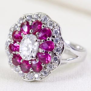 Puede incluir: Un anillo de tono plateado con una piedra central blanca ovalada rodeada por un halo de piedras de color rosa intenso en forma de pera. Un borde exterior festoneado de pequeñas piedras redondas y transparentes enmarca el diseño en una banda dividida.