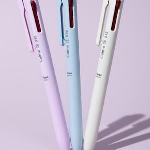May include: Three retractable ballpoint pens in light purple, light blue, and off-white. Each pen features a dark red clip, a textured grip section, and the printed text Calme 3 035 and Pentel BXC33 on the barrel. The pens are arranged vertically.