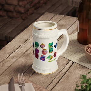 May include: A white ceramic beer stein featuring a gold rim and a handle, decorated with a colourful pattern of various multi-sided gaming dice in shades of green, blue, red, and yellow, with small green text printed near the base of the mug.