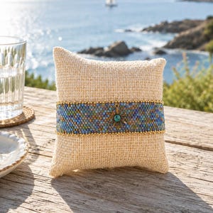 Peut inclure: Un bracelet tissé en perles présentant un motif multicolore de perles bleues, vertes et métalliques. Le bracelet est orné d'un pendentif central en forme de soleil doré vieilli avec une petite pierre turquoise et bordé de perles dorées.
