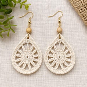 May include: A pair of cream-colored teardrop-shaped crochet earrings featuring a central sunburst pattern. Each earring is suspended from a gold-toned metal hook and accented with a single smooth, light brown wooden bead above the crochet piece.