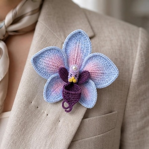 Könnte beinhalten: Eine gehäkelte Orchideen-Brosche mit hellblauen und zartrosa Blütenblättern sowie einem dunkelvioletten Zentrum. Die Blume enthält eine kleine weiße Perle an der Spitze der zentralen Säule und gelbe Details, befestigt an einem beigen, strukturierten Blazer.