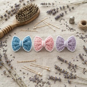 May include: Three crochet hair bows in blue, pink, and light purple, each featuring a white border and a central band. The set includes several gold-toned bobby pins with small white pearl-like beads at the ends, arranged on a textured fabric.