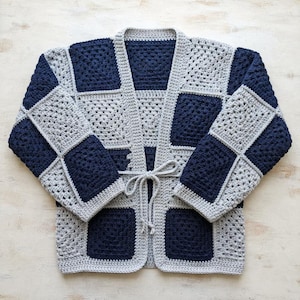 May include: A long-sleeve cardigan made of crocheted granny squares in alternating navy blue and light gray colors. The garment features a front tie closure with matching gray yarn strings and a solid gray crocheted border along the edges.