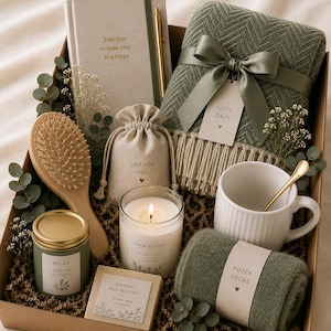Könnte beinhalten: Eine Geschenkbox mit der Aufschrift Ivy Box enthält eine salbeigrüne Fischgrät-Decke mit Fransen, einen weißen gerippten Becher mit goldenem Löffel, ein Notizbuch mit goldenem Stift, eine Sojakerze, eine Holzbürste, Seife und grüne Socken.